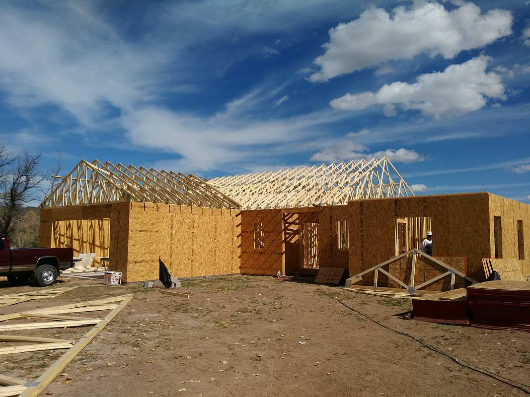 A Tierra Builders residential construction project with timber roof trusses and framed exterior walls beneath a New Mexico sky